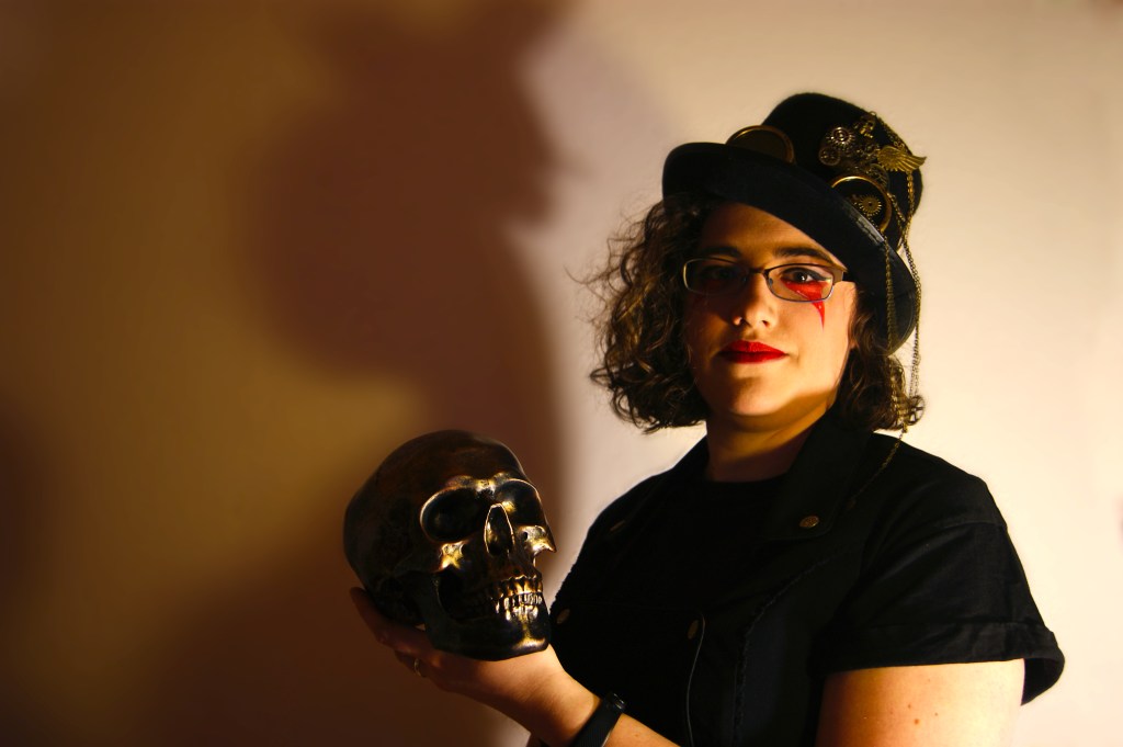 Autorinnenfoto mit einem brozenen Totenkopf in der Hand und einem Zylinder mit Steampunkelementen auf dem Kopf.