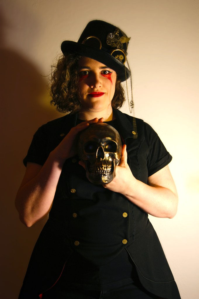 Autorinnenfoto. Ich trage eine schwarze Weste, halte einen bronzenen Totenkopf vor meiner Brust und trage einen schwarzen Zylinder mit Steampunktelementen.